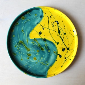 Könnte beinhalten: Ein runder Keramikteller mit einem lebendigen Design. Der Teller ist in zwei Bereiche unterteilt: einen in leuchtendem Gelb und den anderen in Türkis. Schwarze und grüne Farbspritzer fügen dem gelben Bereich Details hinzu. Der Teller ist ein dekoratives Stück.