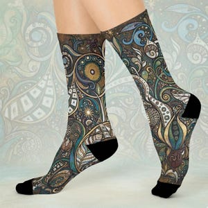 May include: A pair of patterned socks featuring a detailed design in shades of brown, blue, and gold. The socks have black heels and toes, and the intricate pattern includes floral and abstract elements. The socks are displayed against a light blue background.