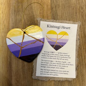 Peut inclure: Ornement en forme de cœur avec une ficelle dorée pour suspendre. L'ornement est peint aux couleurs de la fierté non binaire, avec des lignes dorées ressemblant au kintsugi. Une carte indique "Kintsugi Heart".