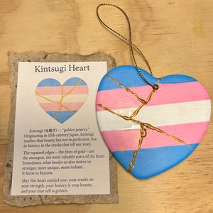 Puede incluir: Adorno en forma de corazón con un diseño a rayas azul, rosa y blanco, con reparaciones de grietas doradas. El adorno va acompañado de una tarjeta con el texto "Kintsugi Heart" y una descripción del arte Kintsugi.