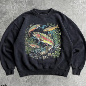 May include: A black crewneck sweatshirt featuring a detailed graphic print of three rainbow trout swimming among green aquatic plants and rocks. The central fish is large and colorful, while two smaller trout are positioned above and below it.