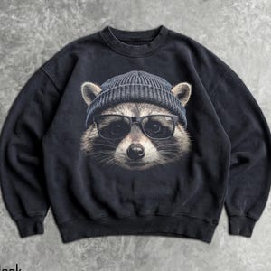 May include: Black sweatshirt featuring a raccoon wearing a blue beanie and black glasses. The raccoon's face is centered on the front of the sweatshirt. The word "Black" is printed at the bottom left corner of the image.