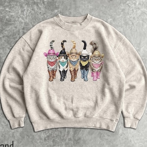 Puede incluir: Sudadera de color arena con un gráfico de cinco gatos vestidos de vaqueros. Cada gato lleva un sombrero de vaquero, botas y un pañuelo o bufanda de varios colores. Los gatos están colocados en fila, creando un diseño juguetón y caprichoso.