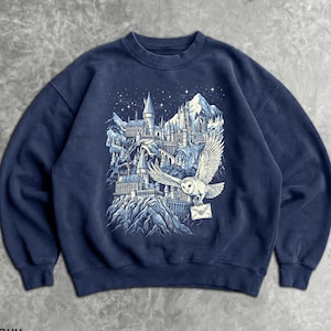 May include: Navy blue sweatshirt featuring a detailed graphic of a castle, mountains, and an owl carrying a letter. The design is in shades of white and blue, set against a starry night sky. The sweatshirt has a crew neck and long sleeves.