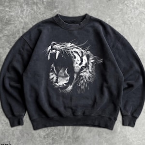 Può includere: Felpa nera girocollo con una grafica dettagliata in bianco e nero di una tigre ruggente. La felpa ha una vestibilità classica con polsini e orlo a coste. La parola "Black" è stampata in basso.