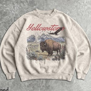 May include: A sand-colored crewneck sweatshirt featuring a graphic print of a bison, wolves, and an eagle in a mountain landscape. The text Yellowstone is printed in red above the illustration, which includes geysers and pine trees.