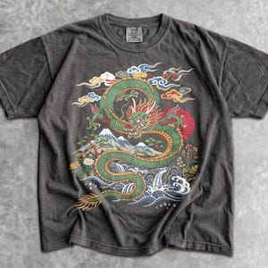 May include: A charcoal gray short-sleeve t-shirt featuring a large graphic print of a green dragon winding through stylized clouds, a snow-capped mountain, and crashing waves. The design includes red, yellow, and blue accents on the dragon.