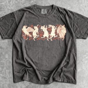 Könnte beinhalten: Ein anthrazitfarbenes Kurzarm-T-Shirt mit einem zentralen Grafikdruck, der eine Krähe, eine Katze, eine Ratte, einen Hasen und eine Fledermaus in einer Reihe inmitten roter Blumenranken zeigt. Das Shirt hat einen Rundhalsausschnitt und ein Label.