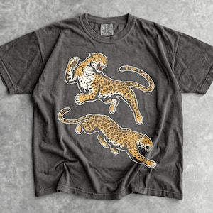 May include: A charcoal gray short-sleeve t-shirt featuring a graphic print of two leaping leopards with yellow, black, and white spotted fur. The shirt has a crew neckline and a faded, washed-out appearance with a visible brand tag at the inner collar.