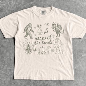 May include: Ivory-colored t-shirt featuring a graphic of various cryptids and the text "respect the locals." The design includes a Bigfoot, an alien, a winged creature, a moth, and the Loch Ness Monster.
