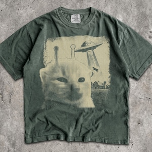 May include: A moss green short-sleeve t-shirt featuring a graphic print of a cat with two antennae on its head. Above the cat, a flying saucer beams down a cow near a farm building. The shirt has a crew neckline and a Comfort Colors brand label.
