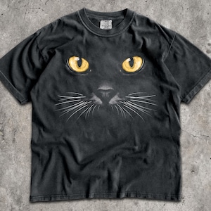 May include: A black short-sleeve t-shirt featuring a graphic print of a black cat face with bright yellow eyes and detailed white whiskers centered on the chest. The shirt has a crew neckline and a Comfort Colors brand label at the inner neck.