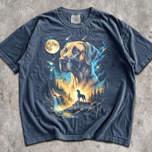 Op de afbeelding: Een marineblauw t-shirt met een grafische print van een grote hondenkop in het bovenste gedeelte, met daaronder een kleiner silhouet van een hond die naar een volle maan huilt boven een boslandschap met een waterval en vallende sterren.