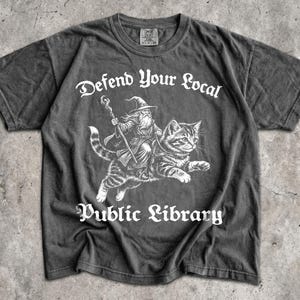 May include: A charcoal grey short-sleeve t-shirt featuring a white graphic of a wizard riding a large cat. The text "Defend Your Local Public Library" is printed in a white gothic font above and below the illustration. The shirt has a crew neckline.
