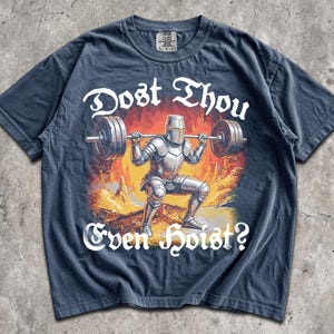 May include: Navy blue t-shirt with a graphic of a knight lifting weights, set against a fiery background. The text "Dost Thou Even Hoist?" is in white script. The shirt is a casual wear item.