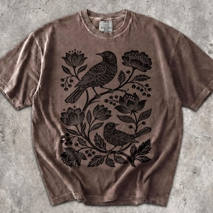 May include: A brown short-sleeve t-shirt featuring a black graphic print of two birds perched among stylized flowers and berries. The shirt has a crew neckline and a label at the inner collar that reads Comfort Colors. The fabric has a washed appearance.