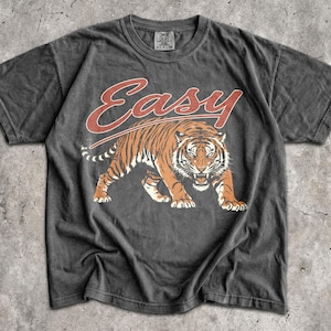 May include: A charcoal grey short-sleeve t-shirt featuring a graphic of a prowling tiger in orange and black tones. Above the tiger, the word "Easy" is printed in a stylized red script font. The shirt has a crew neckline and a faded, worn-in texture.