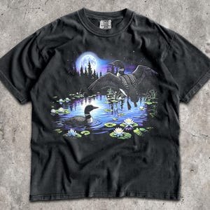 May include: A black short-sleeved t-shirt featuring a graphic print of two loons on a pond at night. One loon is swimming whilst the other is in flight. The scene includes a full moon, a dark forest silhouette, and white water lilies on the water.