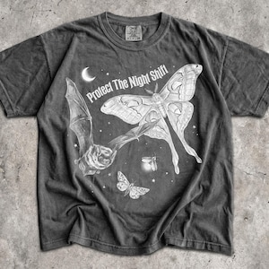 Op de afbeelding: Een antracietgrijs T-shirt met korte mouwen en een witte grafische print van een vleermuis, een grote nachtvlinder, een kleinere vlinder, een maansikkel en sterren. De tekst "Protect The Night Shift" is in het wit boven de afbeelding gedrukt.