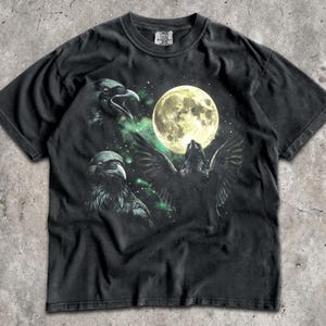 May include: A black short-sleeve t-shirt featuring a graphic print of two ravens and a large full moon against a dark, starry background with green nebula-like accents. The shirt has a crew neckline and a standard fit with a visible brand label.