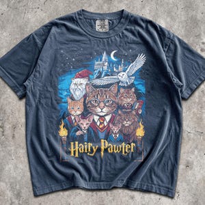 Può includere: T-shirt blu navy con una grafica fantasy di gatti vestiti con abiti da mago, un castello e un gufo. Il testo "Hairy Pawter" è visualizzato sotto.