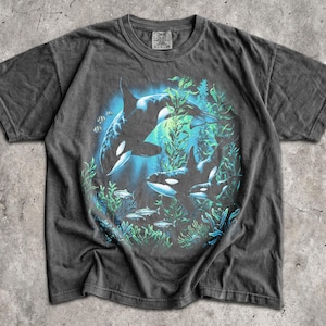 May include: A charcoal gray short-sleeve t-shirt featuring a graphic print of two orcas swimming among green seaweed and small fish. The design is centered on the chest, and the shirt has a crew neckline with a visible brand tag at the collar.