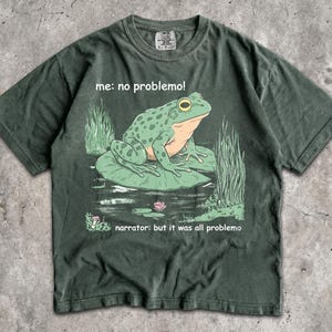 May include: A dark green short-sleeve t-shirt featuring a graphic of a green frog sitting on a lily pad. The text reads "me: no problemo!" above the frog and "narrator: but it was all problemo" below the frog, with reeds and a small flower.