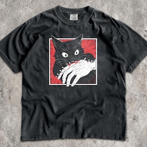 May include: A black short-sleeve t-shirt featuring a graphic print of a black cat biting a human hand. The cat has wide eyes and sharp teeth, set against a red sunburst background within a white square frame. The shirt has a crew neckline.