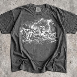 May include: A charcoal gray short-sleeve t-shirt featuring a graphic print of a pack of wolves running through a forest landscape under a stormy sky with bright white lightning bolts. The shirt has a crew neckline and a visible brand label at the neck.