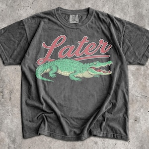 Puede incluir: Camiseta gris oscuro con un dise&ntilde;o de cocodrilo verde y la palabra "Later" en letras rojas. El cocodrilo tiene la boca abierta. La camiseta tiene un aspecto vintage y desgastado.