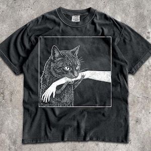 May include: A black short-sleeve t-shirt featuring a white graphic print of a black cat biting a human arm. The design is enclosed within a thin white square border. The shirt has a crew neckline and a faded, washed-out black fabric appearance.