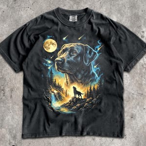 以下が含まれることがあります： 黒の半袖Tシャツ。大きなラブラドールレトリバーの頭部と、滝の近くの岩場に立つ遠吠えする犬のシルエット、満月、流れ星、暗い森が描かれたグラフィックプリントが施されています。