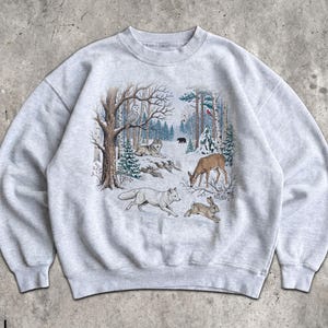 Peut inclure: Un sweat-shirt gris clair à col rond orné d'une illustration détaillée d'une forêt en hiver. Le motif représente un renard blanc, un cerf, un lapin, un hibou et un petit ours noir au milieu d'arbres enneigés et de branches de sapin.