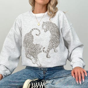 May include: A light gray crewneck sweatshirt featuring a dark gray graphic print of two tigers facing each other in a circular arrangement. The sweatshirt has long sleeves and a ribbed collar, cuffs, and hem. The word Ash is printed in the top left corner.