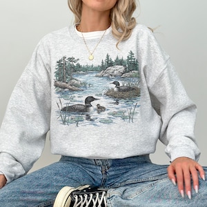 Puede incluir: Una sudadera gris claro de cuello redondo con un estampado gráfico detallado de dos colimbos nadando en un lago con un polluelo, rodeados de pinos y orillas rocosas. La palabra Ash está impresa en la esquina superior izquierda.