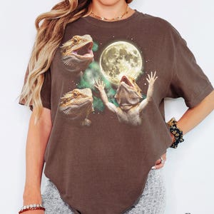 Peut inclure: Un t-shirt à manches courtes marron espresso avec une impression graphique représentant trois dragons barbus. Un lézard tend ses pattes vers une grande pleine lune brillante entourée de nuages nébuleux verts et d'étoiles blanches.