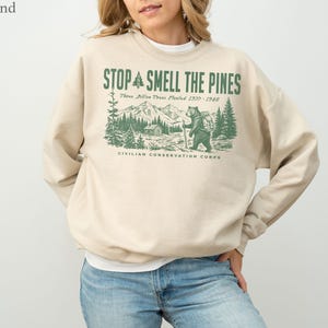 Pode incluir: Uma camisola de gola redonda cor de areia com uma impress&atilde;o gr&aacute;fica verde de um urso a caminhar numa floresta com montanhas e uma cabana. O texto diz STOP SMELL THE PINES, Three Billion Trees Planted 1977-1942, CIVILIAN CONSERVATION CORPS.