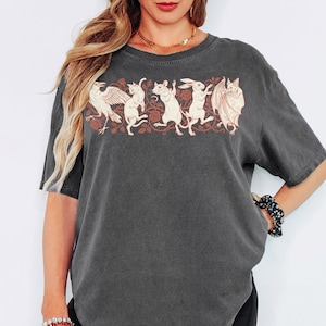 Puede incluir: Una camiseta de manga corta en color gris carbón con un estampado gráfico de un cuervo, un gato, un ratón, un conejo y un murciélago alineados entre enredaderas florales. La ilustración está representada en un tono crema claro.