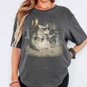Könnte beinhalten: Ein anthrazitfarbenes Kurzarm-T-Shirt mit einem Grafikdruck einer Ratte, die eine schwarze Sonnenbrille trägt und kleine Hanteln in einem Fitnessstudio hebt, mit einer klaren Wasserflasche auf dem Boden und dem Wort Pepper.