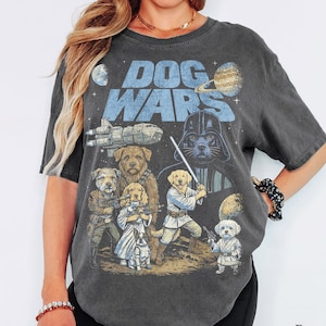 Może przedstawiać: Ciemnoszary t-shirt z krótkim rękawem z nadrukiem graficznym i napisem DOG WARS. Wzór przedstawia różne rasy psów przebrane za postacie z science fiction, w tym psa w ciemnym hełmie trzymającego świecący miecz.