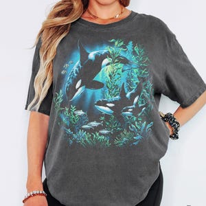 Puede incluir: Una camiseta de manga corta gris carbón con un estampado gráfico de dos orcas nadando entre algas verdes y peces pequeños. La camiseta tiene cuello redondo y un corte holgado. La palabra Pepper está impresa en la esquina inferior derecha.