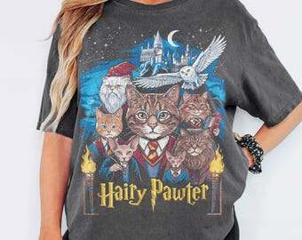 Puede incluir: Una camiseta gris carbón de manga corta con un estampado gráfico de varios gatos vestidos con túnicas de mago. El diseño incluye un castillo, un búho volando y el texto Hairy Pawter en letras amarillas. Los gatos tienen diversos pelajes.