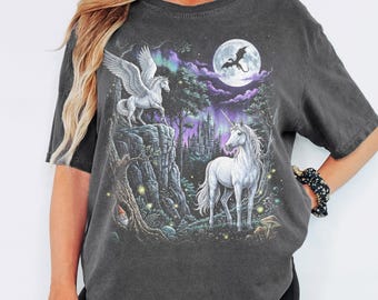 Puede incluir: Una camiseta gris carbón de manga corta con un estampado gráfico de un pegaso blanco sobre un acantilado, un unicornio blanco en un bosque, un castillo lejano, una luna llena con la silueta de un dragón volador y auroras boreales moradas.