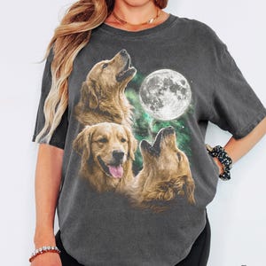 以下が含まれることがあります： ダークグレーの半袖Tシャツ。暗く霧がかった背景に浮かぶ大きく明るい満月を見上げる3匹のゴールデンレトリバーのグラフィックプリントが施されています。クルーネックでゆったりとしたフィット感のTシャツです。