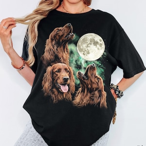 Op de afbeelding: Een zwart T-shirt met korte mouwen en een grafische print van drie bruine Ierse setters. Twee honden kijken met open bek omhoog, terwijl de middelste hond met de tong uit de bek naar voren kijkt. Een grote, heldere volle maan schijnt erboven.
