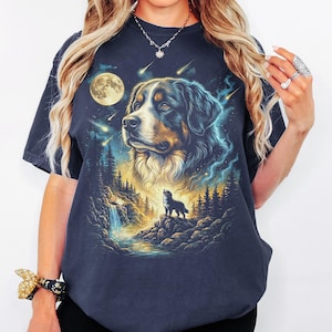 May include: A navy blue short-sleeve t-shirt featuring a graphic print of a Bernese Mountain Dog head above a smaller silhouette of a dog standing on a rocky cliff near a waterfall, set against a night sky with a full moon and falling stars.