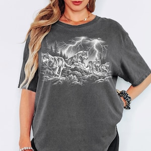 May include: A charcoal gray short-sleeve t-shirt featuring a white graphic print of a pack of wolves running through a forest landscape under a stormy sky with lightning bolts. The shirt has a crew neckline and a relaxed, loose-fitting silhouette.