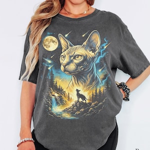 May include: A charcoal grey short-sleeve t-shirt featuring a graphic print of a Sphynx cat head above a smaller cat silhouette standing on a rocky cliff near a waterfall, set against a night sky with a full moon, falling stars, and trees.