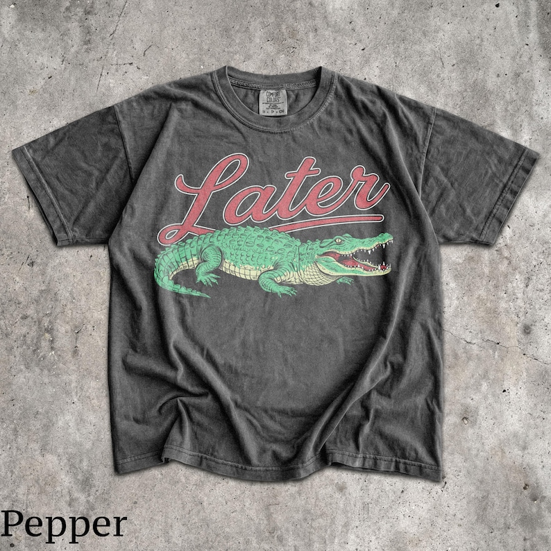 Puede incluir: Camiseta gris oscuro con un dise&ntilde;o de cocodrilo verde y la palabra "Later" en letras rojas. El cocodrilo tiene la boca abierta. La camiseta tiene un aspecto vintage y desgastado.