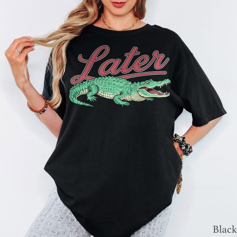 May include: A black short-sleeved t-shirt featuring a graphic of a green alligator positioned below the word "Later" written in a red cursive font. The design is centered on the chest of the garment, which has a relaxed fit and crew neckline.